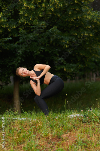 Full length portrait of a young woman in black sportwear in the park doing yoga in the park, trees background. Vertical view. © Dragosh
