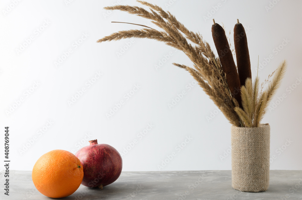 An orange and pomegranate on a textured gray table against a white wall with an autumn bouquet of dried herbs and reeds.