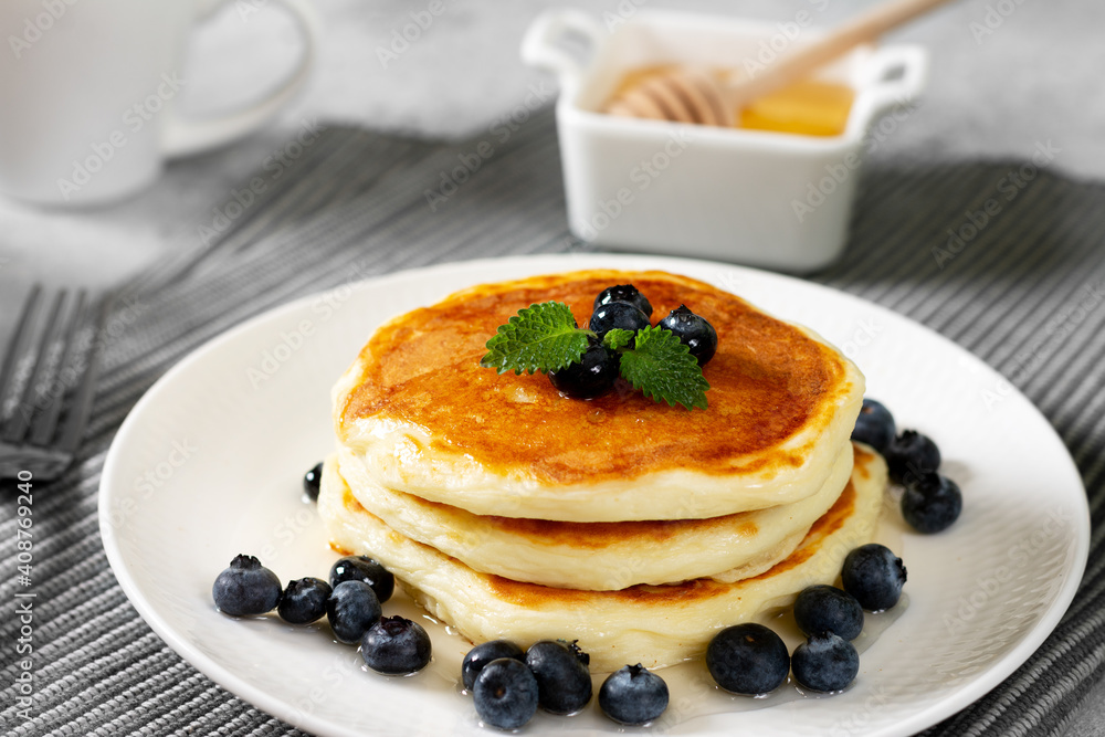 Pancakes in a white plate on the light gray kitchen table. Homemade delicious pancakes with honey and blueberries. Delicious sweet and hearty breakfast Pancakes in a white plate on the light gray kitchen table. Homemade delicious pancakes with honey and blueberries. Delicious sweet and hearty breakfast