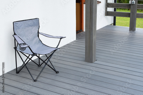 Part of white residential house with dark gray or anthracite wpc composite material terrace and empty chair on deck with wooden railings outdoors.
