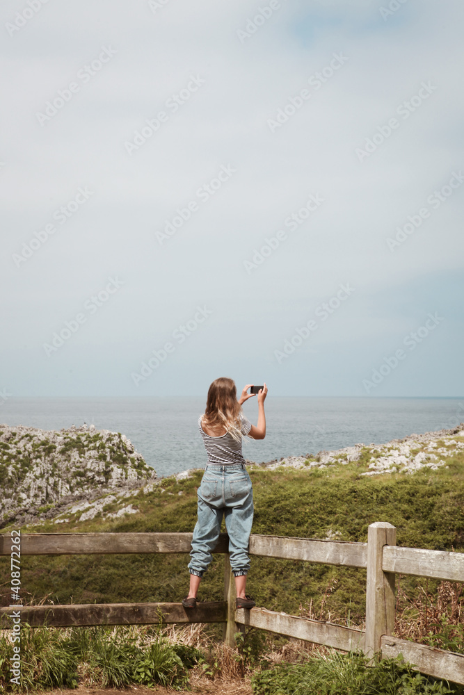 young woman taking photography of the ocean coastline with her phone, standing on the wooden barrier on the cliff