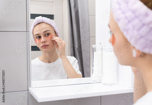 Skin care concept. Young girl puts patches on her face in front of the bathroom mirror