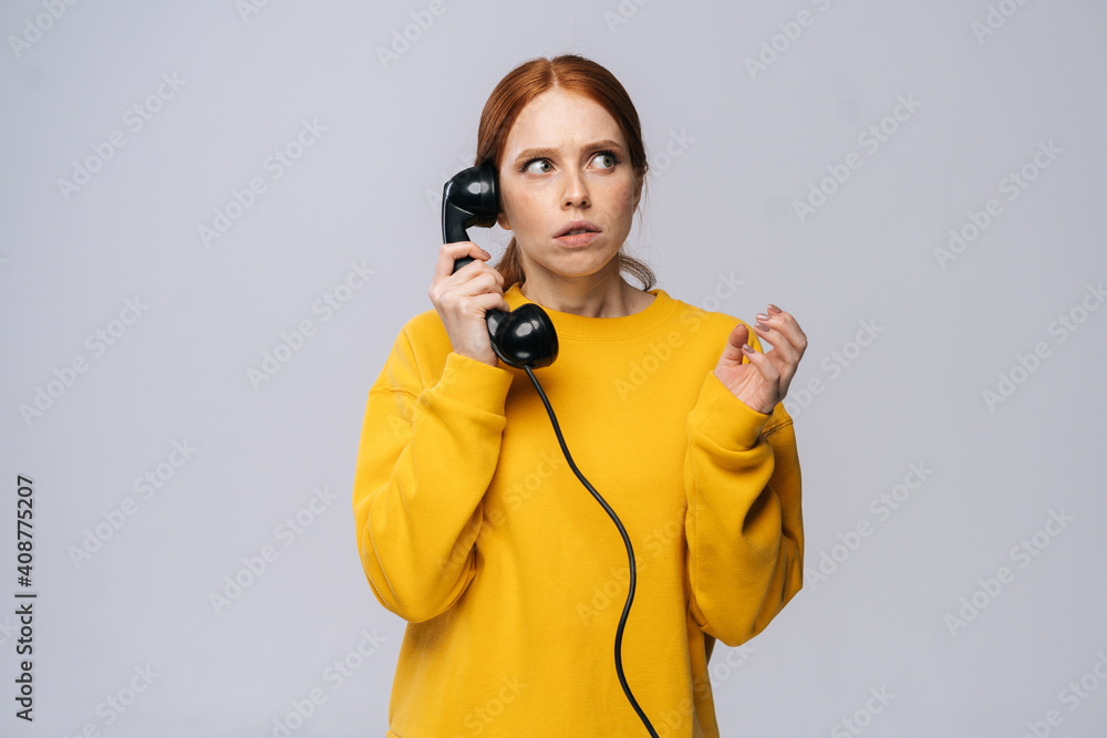 Terrified young woman wearing stylish yellow sweater talking on retro phone looking away against isolated white background. Pretty redhead lady model emotionally showing facial expressions. Terrified young woman wearing stylish yellow sweater talking on retro phone looking away against isolated white background. Pretty redhead lady model emotionally showing facial expressions.