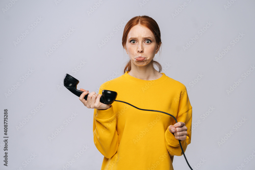 Confused young woman in stylish yellow sweater holding handset receiver of retro phone and looking at camera against white background. Pretty redhead lady model emotionally showing facial expressions. Confused young woman in stylish yellow sweater holding handset receiver of retro phone and looking at camera against white background. Pretty redhead lady model emotionally showing facial expressions.