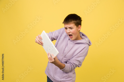 Playing tablet. Caucasian girl's portrait isolated on yellow background with copyspace. Looks excitable. Beautiful female model in hoodie. Concept of human emotions, facial expression, sales, ad