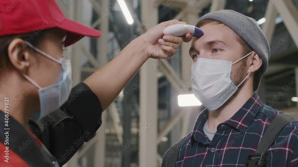 Slow-motion close-up of female foreman measuring worker temperature with contactless thermometer standing at factory, both wearing protective masks Slow-motion close-up of female foreman measuring worker temperature with contactless thermometer standing at factory, both wearing protective masks