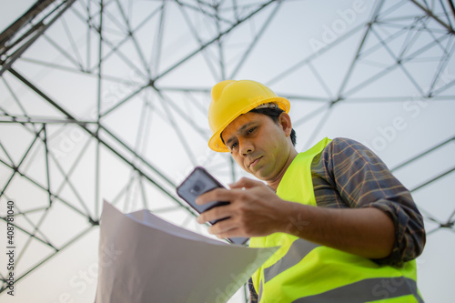 Electrical engineers asian holding blueprint and using smartphone inspect the high voltage pole