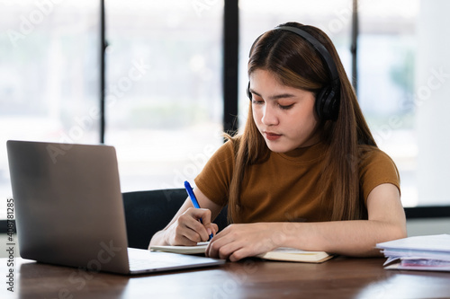 Young Asian girl student wears wireless headphones concentrates write on the notebook to study language online watch and listen to the lecturer, webinar via video call e-learning at home