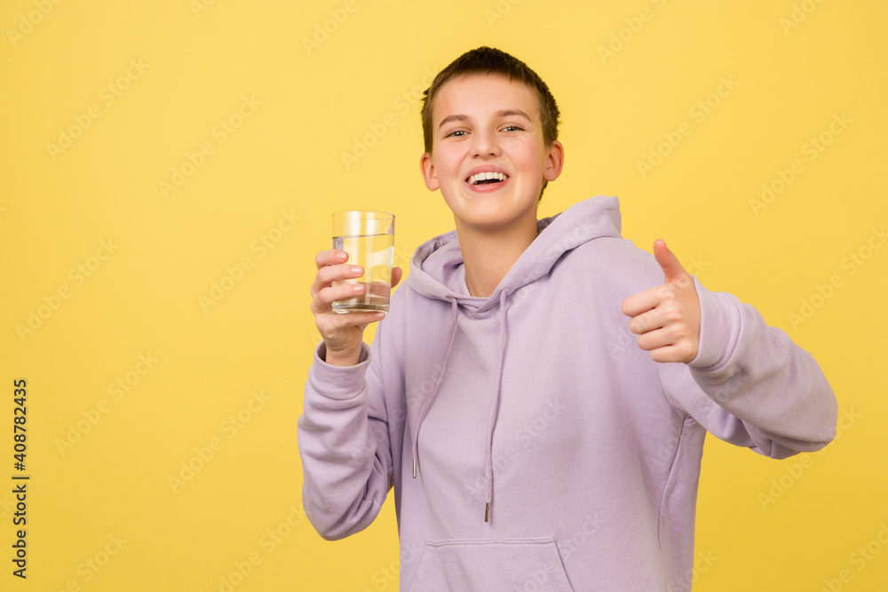 Drinks water, thumb up. Caucasian girl's portrait isolated on yellow background with copyspace. Beautiful female model in hoodie. Concept of human emotions, facial expression, sales, ad, fashion. Drinks water, thumb up. Caucasian girl's portrait isolated on yellow background with copyspace. Beautiful female model in hoodie. Concept of human emotions, facial expression, sales, ad, fashion.
