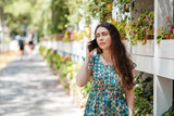 A long-haired woman in a summer dress and a straw hat is talking on the phone. Park street in the background. Concept of communication and social networks