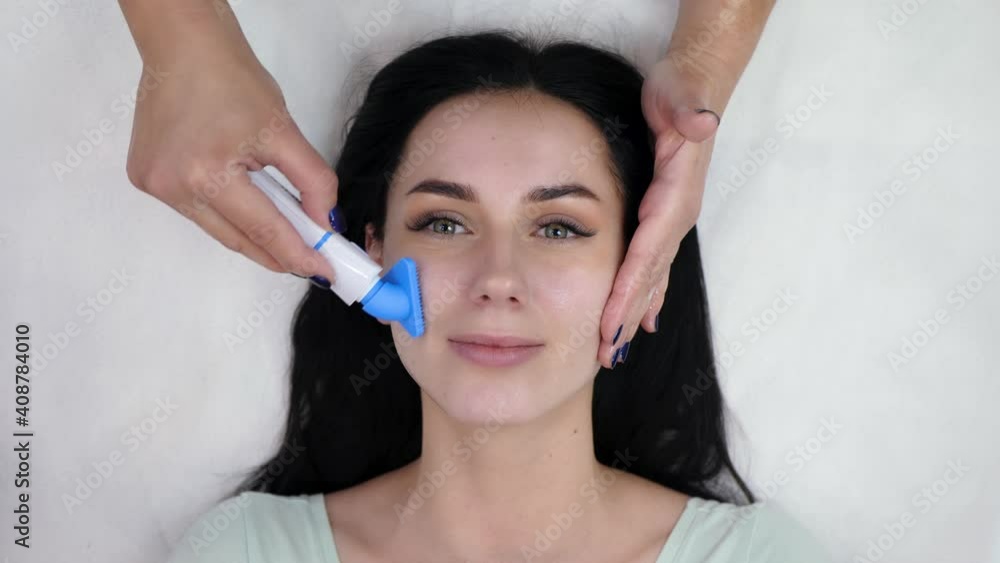 Beautician washes the woman's face with a special cleaning vibrating brush top view. Beauty woman with green eyes looking at camera, isolated on white background. Preparing the skin for the procedure