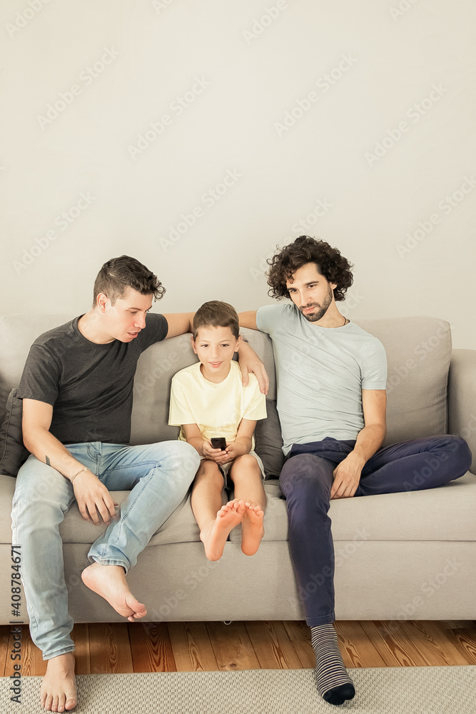 Two dads and son watching TV show at home, sitting on couch in living room, hugging, talking, looking away. Family and home entertainment concept Two dads and son watching TV show at home, sitting on couch in living room, hugging, talking, looking away. Family and home entertainment concept