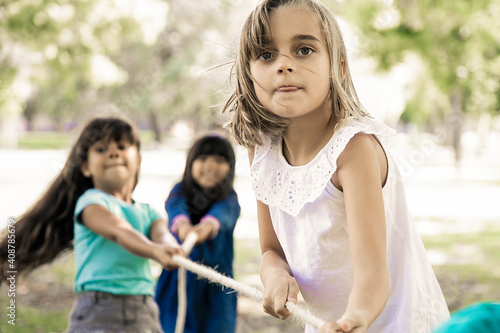 Excited girl enjoying outdoor activities with classmates, playing tug-of-war with friends. Group of children having fun in park. Childhood concept