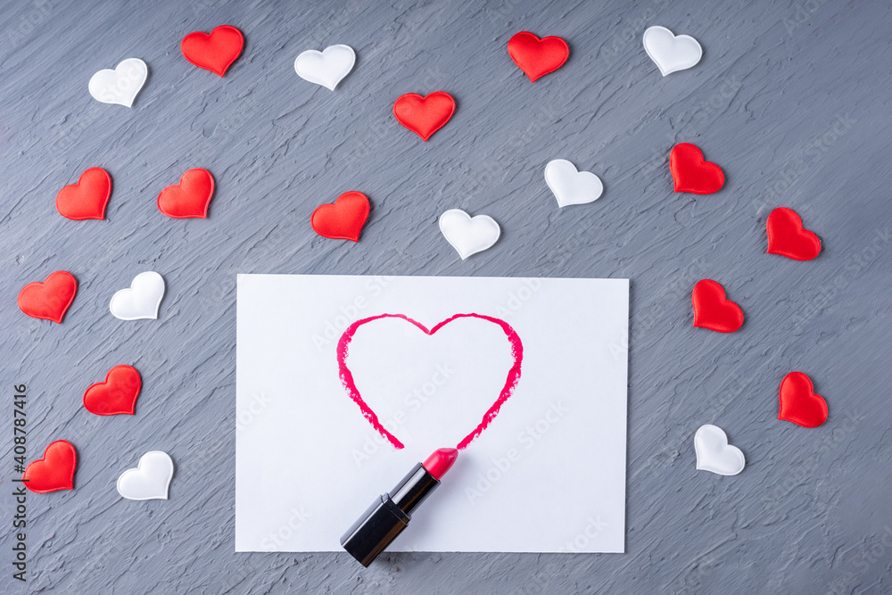 Fototapeta premium Lipstick draws a heart shape on white paper on a gray background with white and red hearts, copy space, flat lay, top view, flat lay. Valentines day background