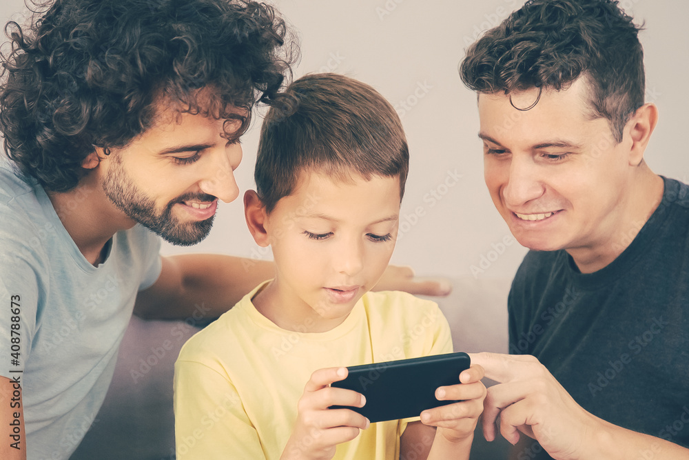 Two happy dads teaching son to use online app on cell. Boy playing game on mobile phone. Family at home and communication concept