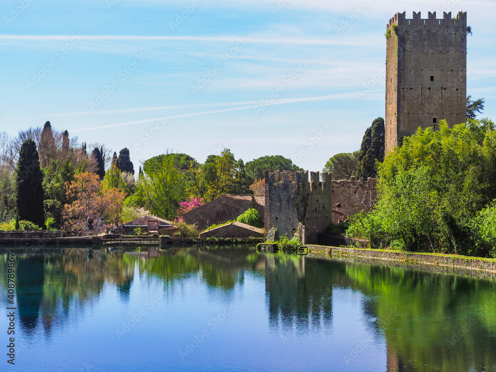 Fototapeta premium Botanical park, Garden of Ninfa or Giardino di Ninfa near the lake, with medieval castle ruins of Caetani family and various plants. Amazing scenery during spring. Beautiful landscape with blue sky.
