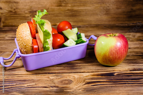 Ripe apple and lunch box with hamburger, cucumbers and tomatoes on wooden table