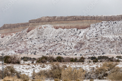 Red rock mountain with slight snow cover behind brush filled desert vista landscape in rural New Mexico on overcast day
