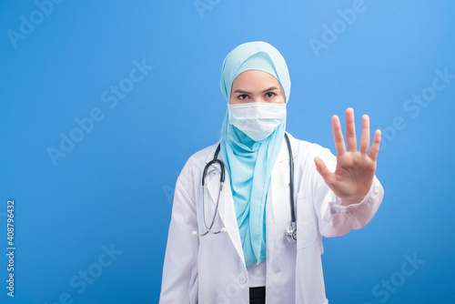 Female muslim doctor with hijab wearing a surgical mask over blue background studio.