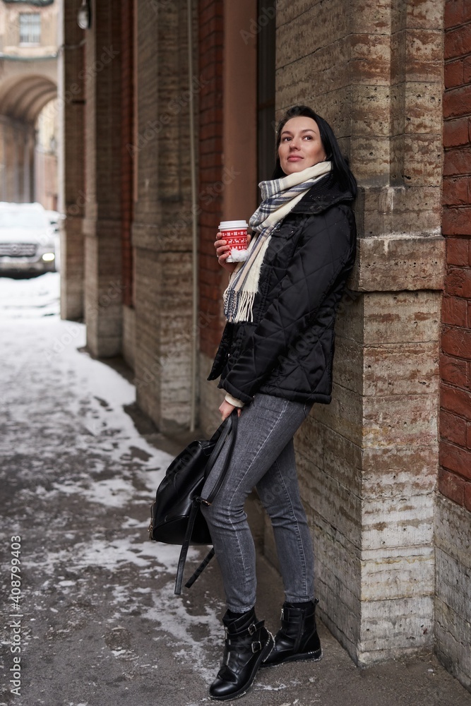 a beautiful girl with shoulder-length dark hair stands in winter clothes, leaning her back on a stone wall and holding a red disposable cup in her hands a beautiful girl with shoulder-length dark hair stands in winter clothes, leaning her back on a stone wall and holding a red disposable cup in her hands