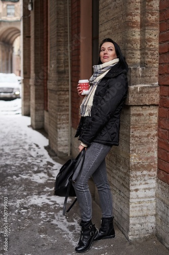 a beautiful girl with shoulder-length dark hair stands in winter clothes, leaning her back on a stone wall and holding a red disposable cup in her hands © Elena