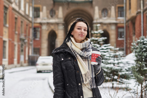 a beautiful girl with shoulder-length dark hair stands against the background of snow-covered trees and a beautiful building and holds a glass of drink in her hands