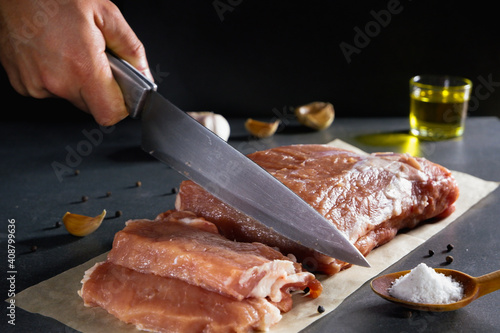 Cooking. Male hands with a knife cut a large piece of raw fresh meat on a stone gray background next to spices