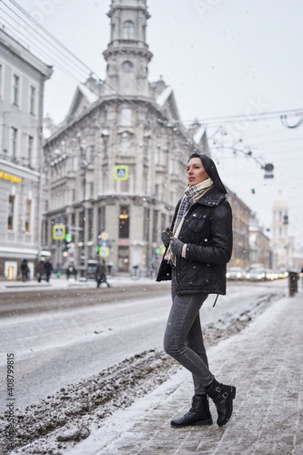 a beautiful girl with shoulder-length dark hair stands in the street in dark outerwear. snowing