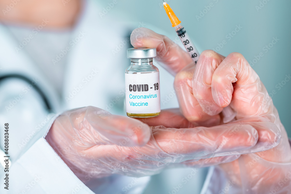 Doctor or nurse in uniform and gloves wearing face mask protective in lab holding medicine vial vaccine bottle with COVID-19 Coronovirus vaccine label Doctor or nurse in uniform and gloves wearing face mask protective in lab holding medicine vial vaccine bottle with COVID-19 Coronovirus vaccine label