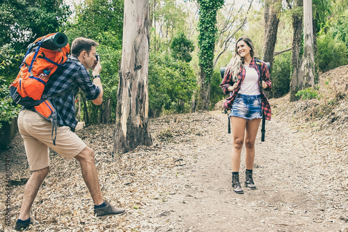 Blonde woman posing for photo on road in forest. Caucasian man holding camera and shooting on nature. Two happy people trekking with backpacks. Tourism, adventure and summer vacation concept © Mangostar