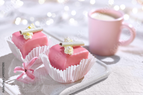 Valentine petit fours with marzipan icing and cream flowers. Espresso coffee in pink cup. Happy Valentine's day Garland of lights on ivory, off white textile tablecloth. Pink hyacinth flower.