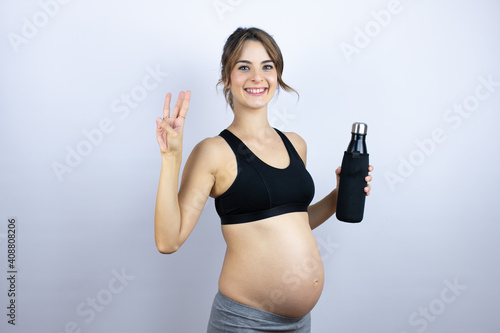Young sportswoman pregnant wearing sportswear holding bottle with water over white background showing and pointing up with fingers number three while smiling confident and happy © Irene