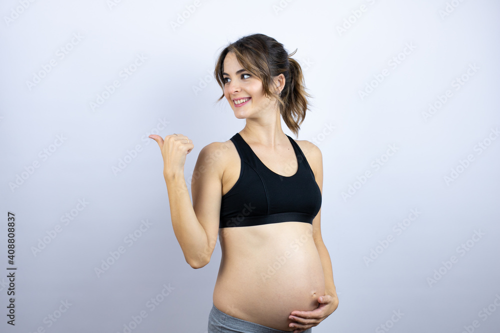 Young sportswoman pregnant wearing sportswear over white background smiling and pointing with hand and finger to the side Young sportswoman pregnant wearing sportswear over white background smiling and pointing with hand and finger to the side