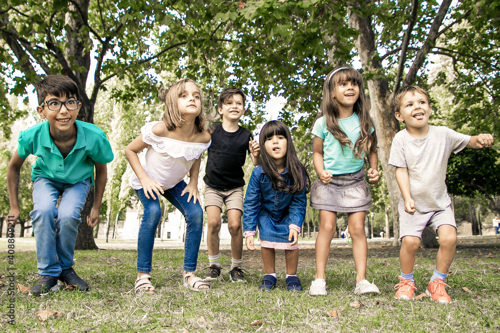 Row of cheerful kids doing squats together in park, looking away in excitement. Kids party or entertainment concept Row of cheerful kids doing squats together in park, looking away in excitement. Kids party or entertainment concept