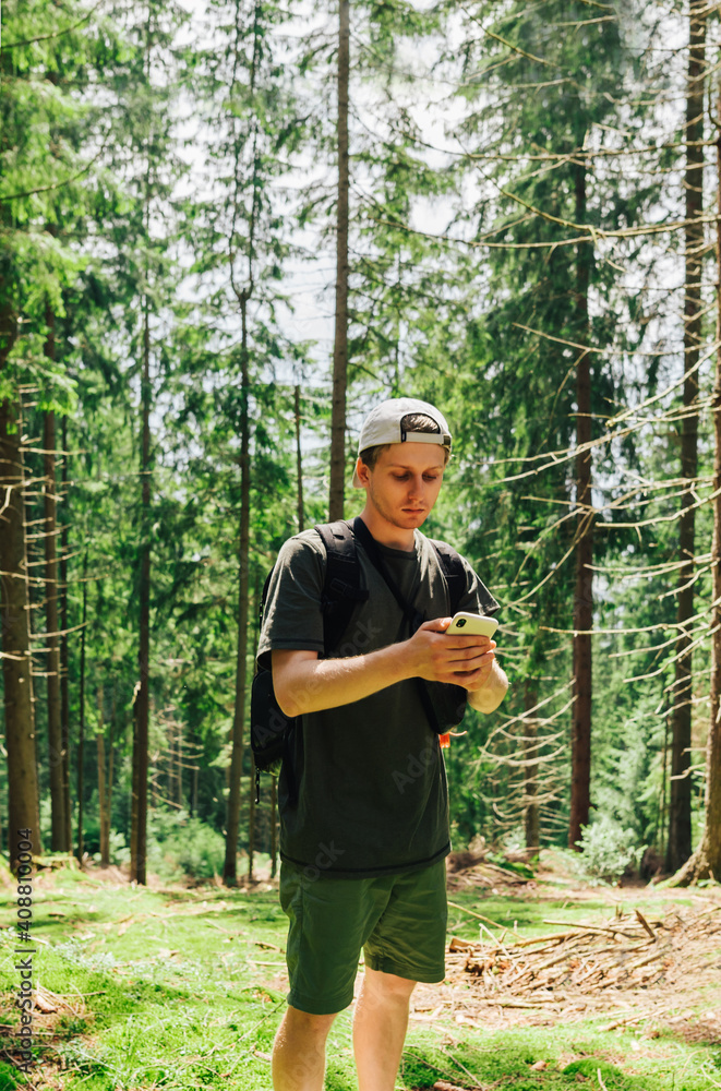 Handsome young man hiker in casual clothes uses a smartphone in a thick coniferous forest. Vertical. Handsome young man hiker in casual clothes uses a smartphone in a thick coniferous forest. Vertical.