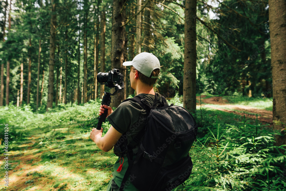 Fototapeta premium Young male hiker in casual clothes stands in a mountain forest with a camera on a stabilizer and makes a video. Video operator captures content on camera during th