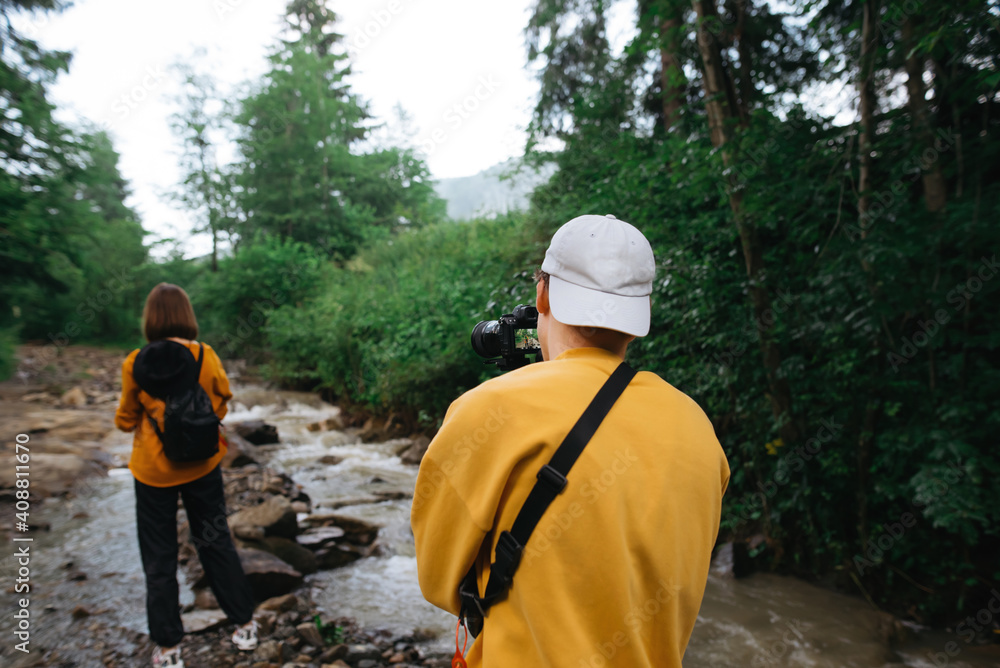 Young man with a camera on a stabilizer shoots a woman on video in the mountains near a stream. Cameraman shoots travel video on camera with a friend. Young man with a camera on a stabilizer shoots a woman on video in the mountains near a stream. Cameraman shoots travel video on camera with a friend.