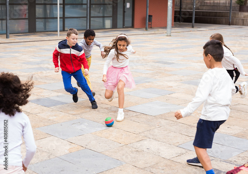 Happy tween girls and boys of different nationalities playing football in schoolyard during break in lessons on warm fall day.