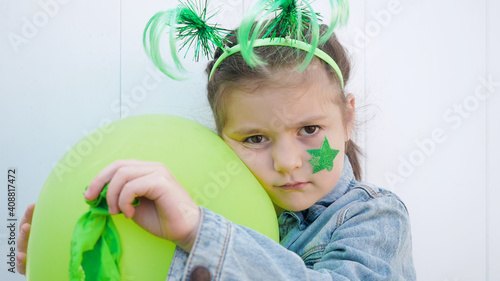 Upset girl with green funny horns on head and star on cheek huging green balloon and making sad face looking at the camera, celebrating saint patrick's day. White wall background