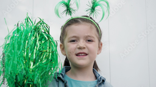 Cute happy brown hair child with green funny horns playing with green pom poms white wall background, celebrating saint patrick's day.