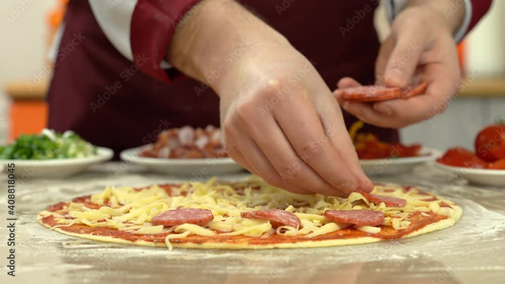 Cooking pizza in kitchen. Chef puts thin slices of sausage on pizza dough. Professional baker prepares traditional homemade Italian pizza. Ingredients near pizza. Cooking pizza in kitchen. Chef puts thin slices of sausage on pizza dough. Professional baker prepares traditional homemade Italian pizza. Ingredients near pizza.