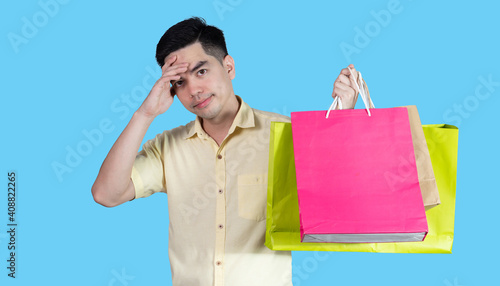 Portrait handsome young asian man wearing yellow stressful content guy carrying new things colorful packages on blue background in studio. Asian man people. photo