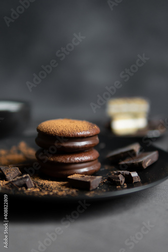Round chocolate brownie on a black plate and cocoa powder on a gray concrete table. Sweets and coffee background.