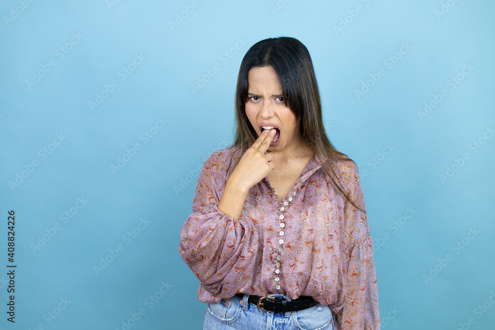 Beautiful latin woman wearing a pink shirt over blue isolated background disgusted with her hand inside her mouth Beautiful latin woman wearing a pink shirt over blue isolated background disgusted with her hand inside her mouth