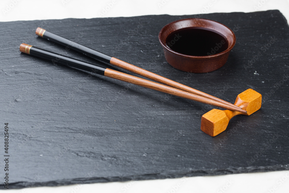 .Black board on a white background. Empty black granite stone rectangular board on white textured background. sushi sticks and soy sauce on coasters on a stone board .Black board on a white background. Empty black granite stone rectangular board on white textured background. sushi sticks and soy sauce on coasters on a stone board