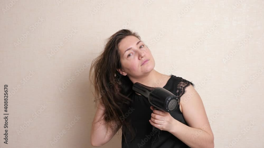 Nice and calm lady in black t-shirt enjoys drying long hair with modern styler standing against white wall in room close-up Nice and calm lady in black t-shirt enjoys drying long hair with modern styler standing against white wall in room close-up