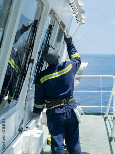 Deck worker, ordinary seaman in blue overalls painting with brush the bridge in vessel to white color.