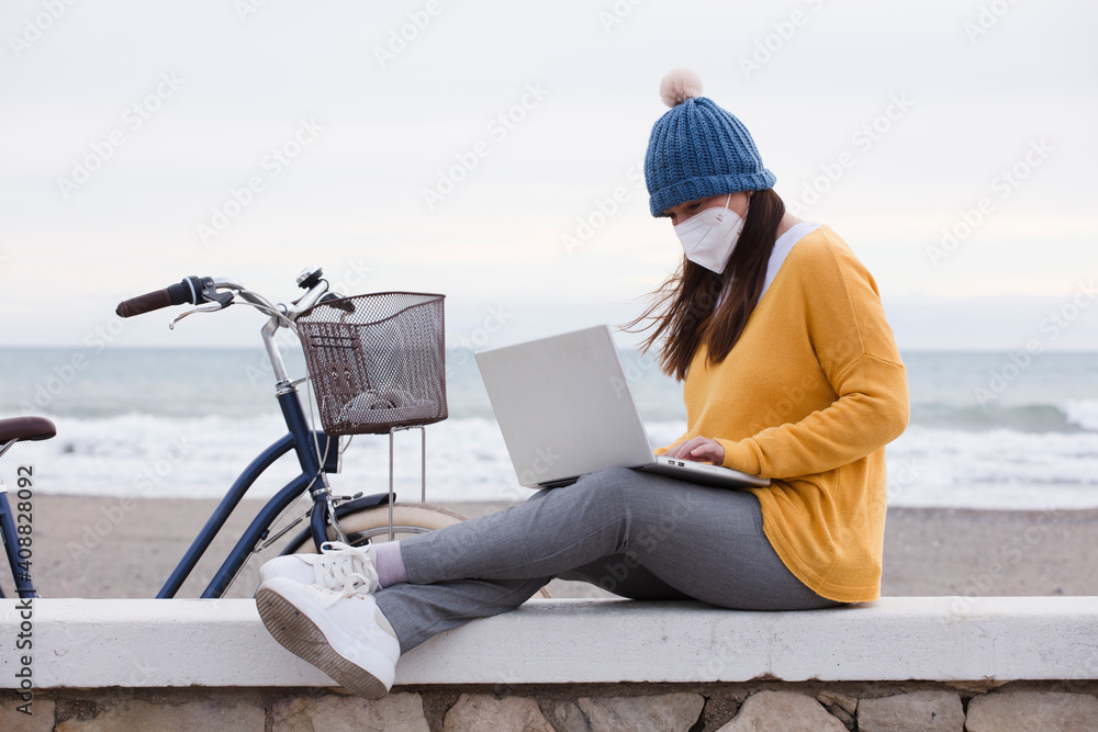 Photo of a woman with a mask, freelancer works outdoors on a laptop connected to public wifi. working online while spending time by the sea