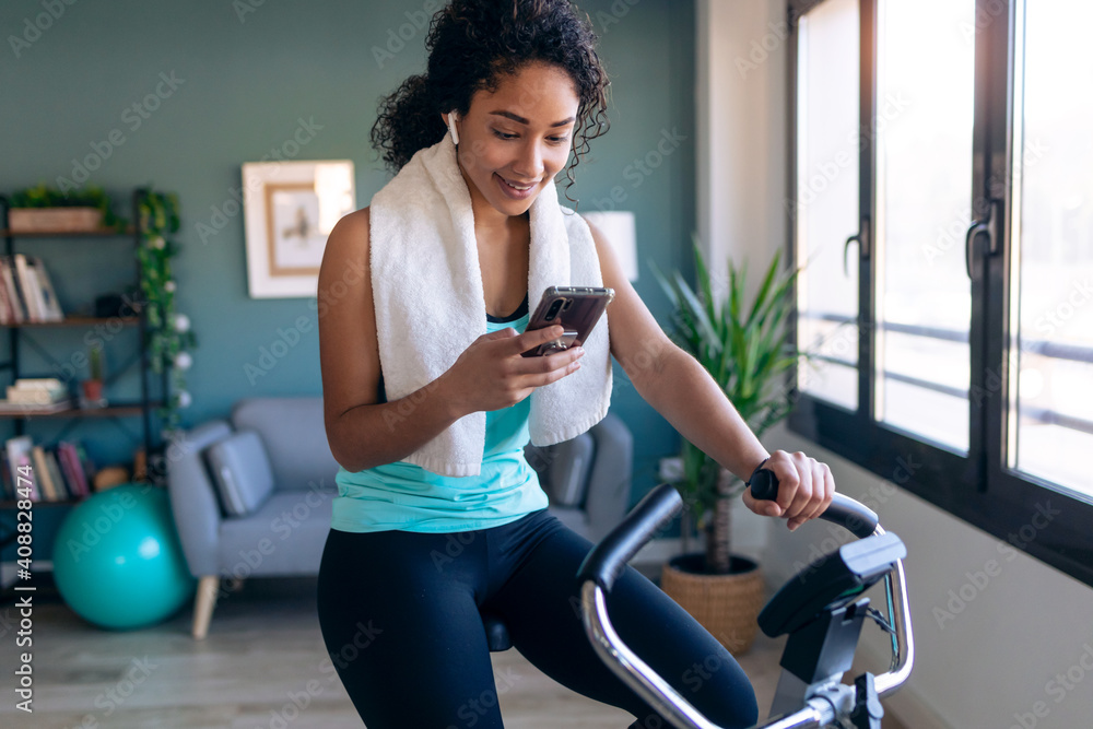 © nenetus - Afro young fitness girl using mobile phone while training on exercise bike at home. © nenetus - Afro young fitness girl using mobile phone while training on exercise bike at home.