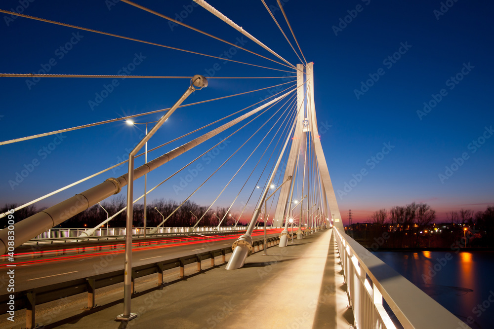 Evening cityscape with long exposure. Bridge over the river, illuminated by lanterns and passing cars in the background of the sunset.